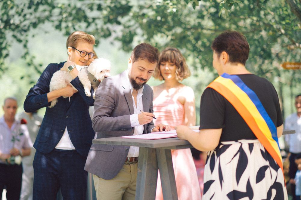 Wedding guests gather around signing table while holding small white dog during outdoor celebration.