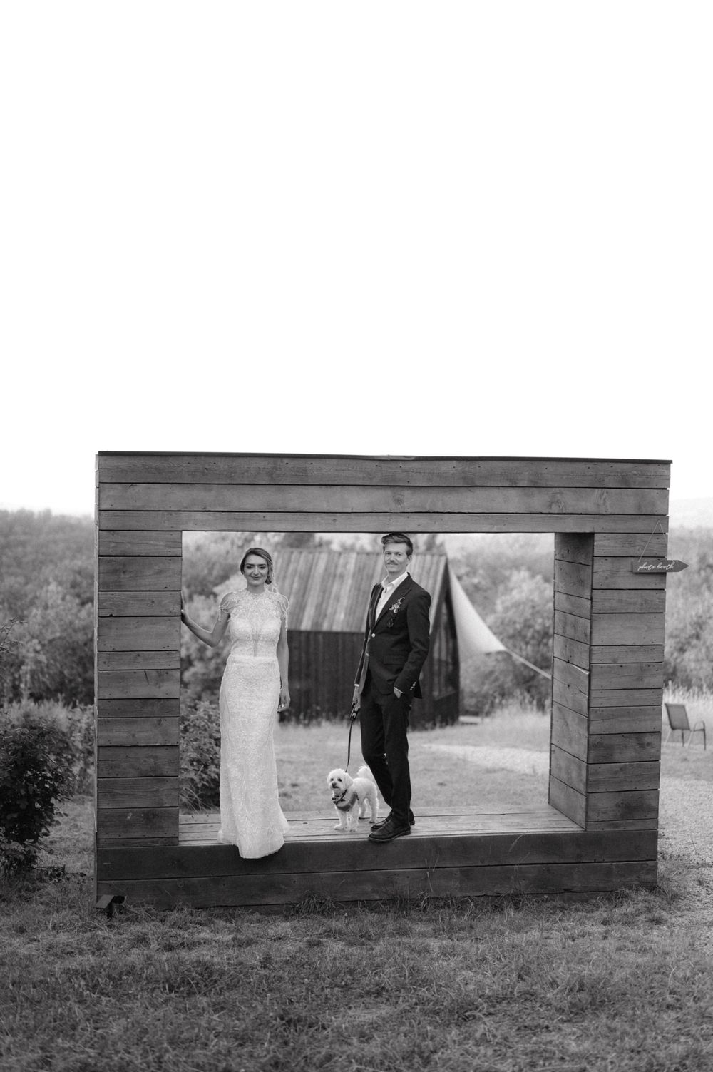 Rustic wedding photo through wooden frame archway with couple and small dog in black and white.