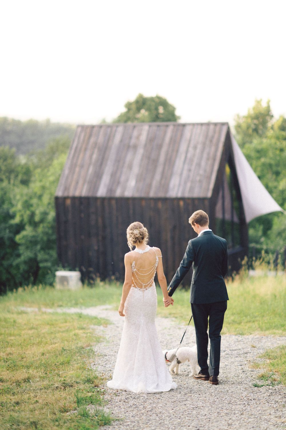 Bride and groom walking hand in hand with small white dog on path near rustic barn.