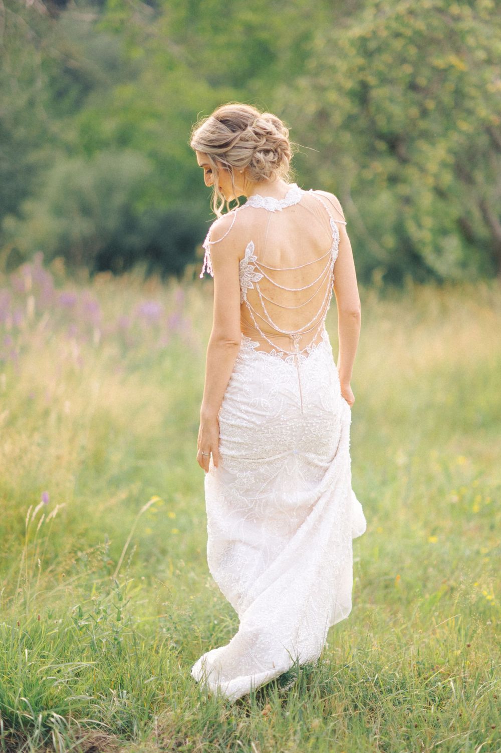 Back view of wedding dress showing intricate lace and chain details against meadow backdrop.