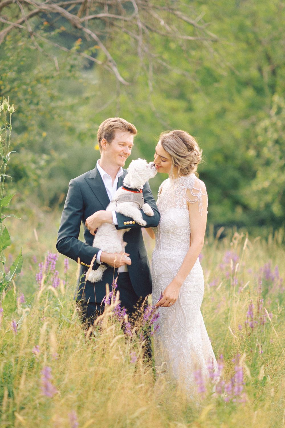 Wedding couple standing in wildflower meadow holding small white dog.