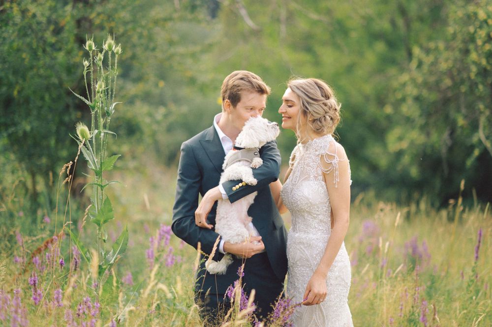 Romantic wedding portrait in field of purple wildflowers with couple holding small dog.