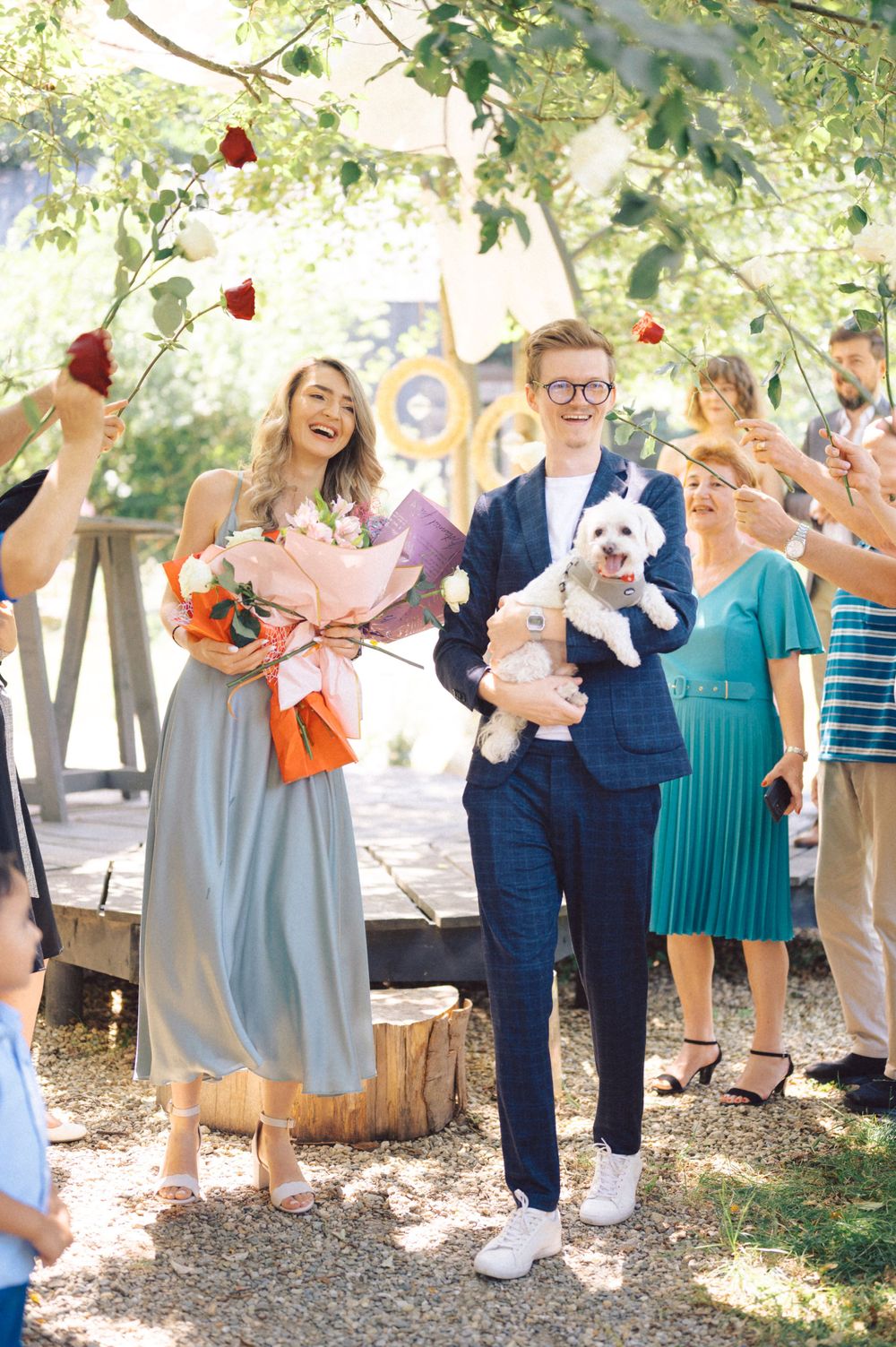 Outdoor celebration with guests throwing rose petals as couple holds bouquet and small white dog.