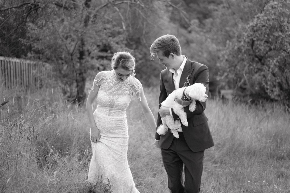 Black and white candid wedding photo of couple laughing while holding small white dog.