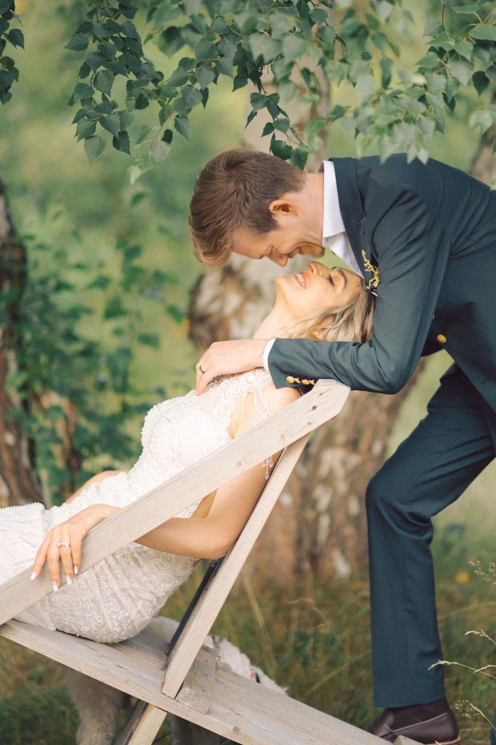 Newlyweds share a tender moment on a wooden bench surrounded by green leaves.