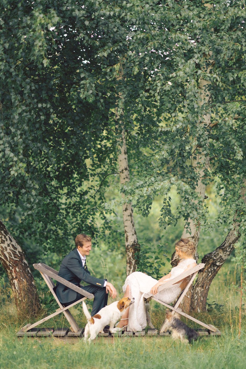 Bride and groom sitting in wooden chairs in garden with pets.