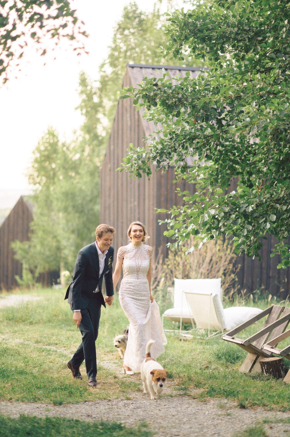 Couple walking with dog on path beside rustic wooden buildings.