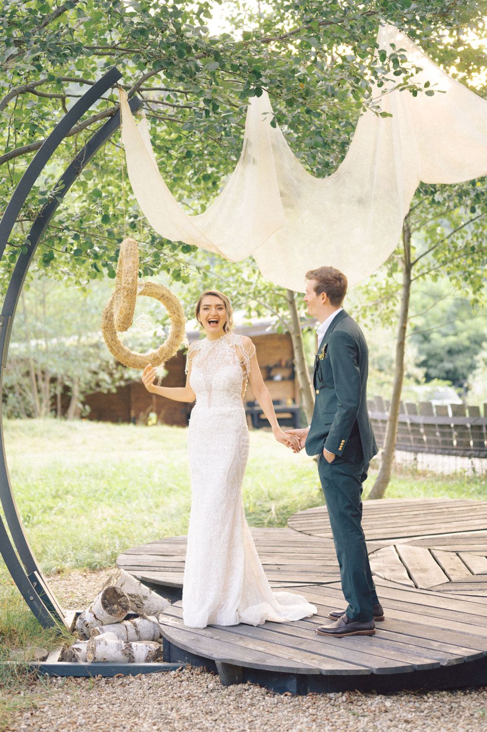 A couple stands beneath white fabric draping and greenery on a wooden deck.