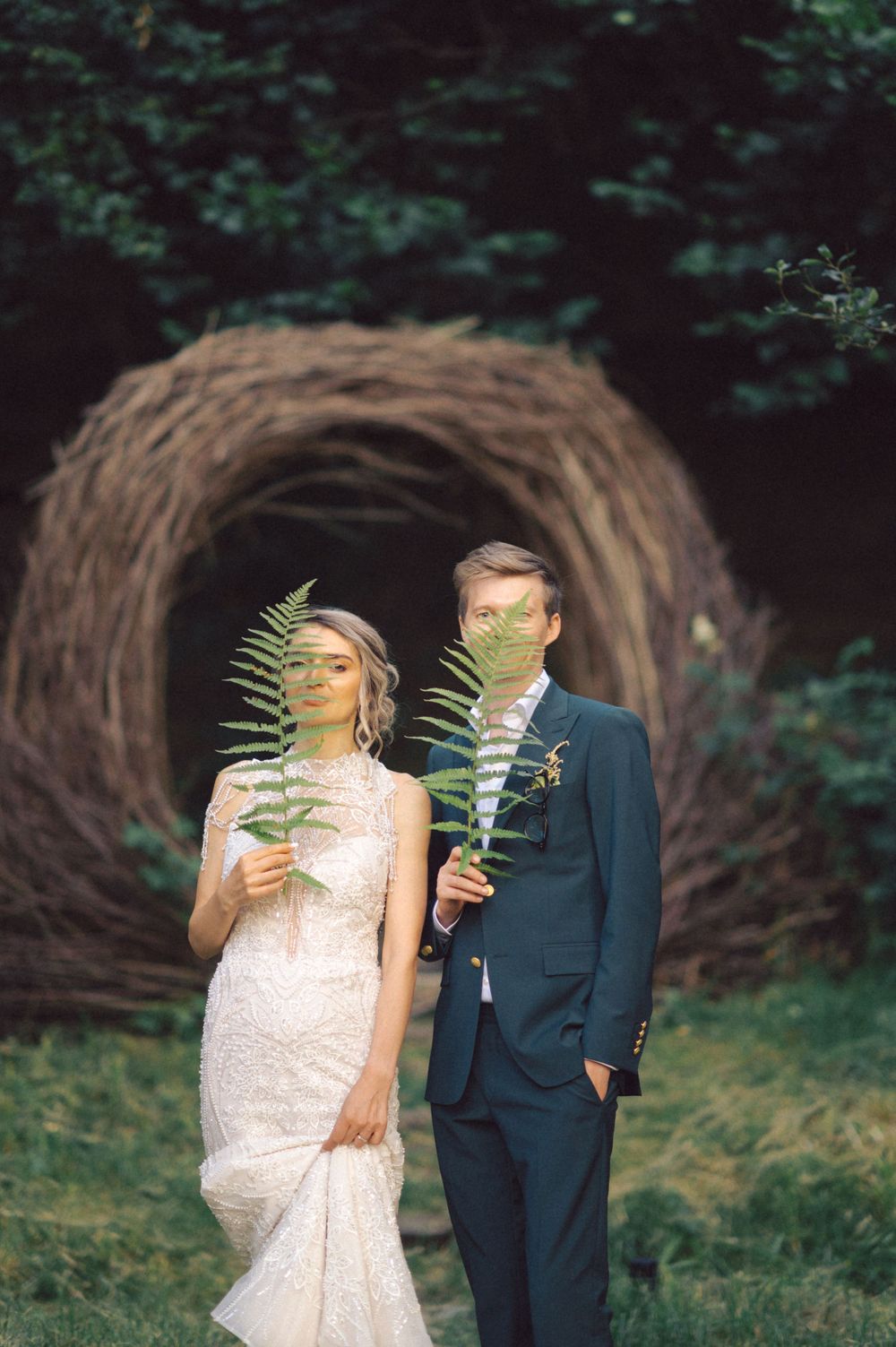 A couple holding fern leaves poses in front of a large woven nest structure.