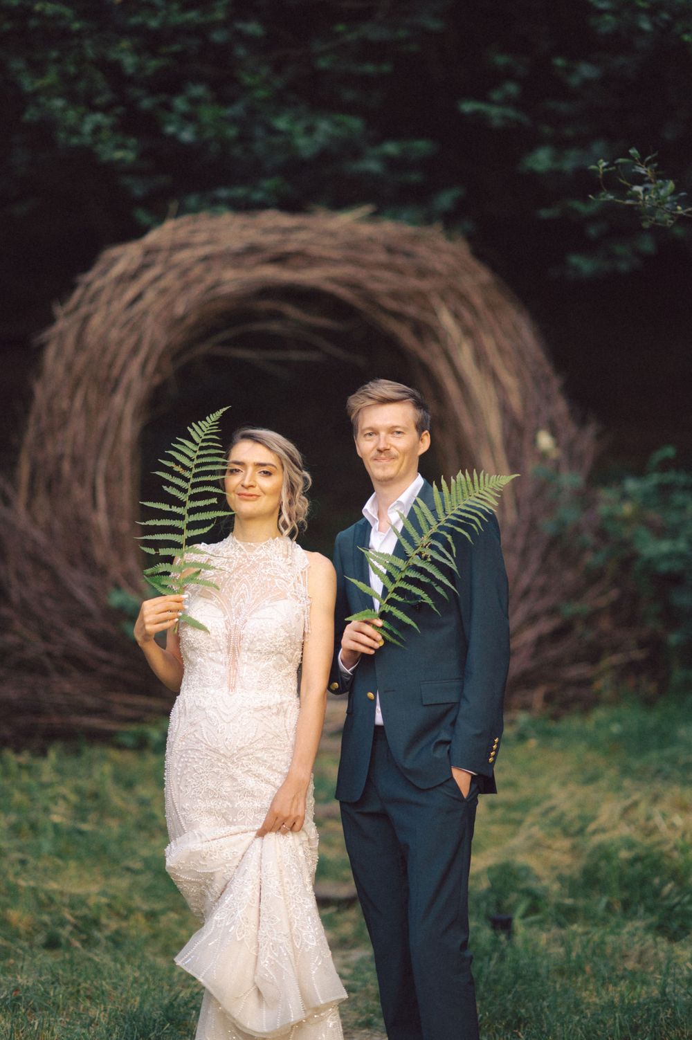 Newlyweds pose with fern fronds in front of a natural woven tunnel structure.