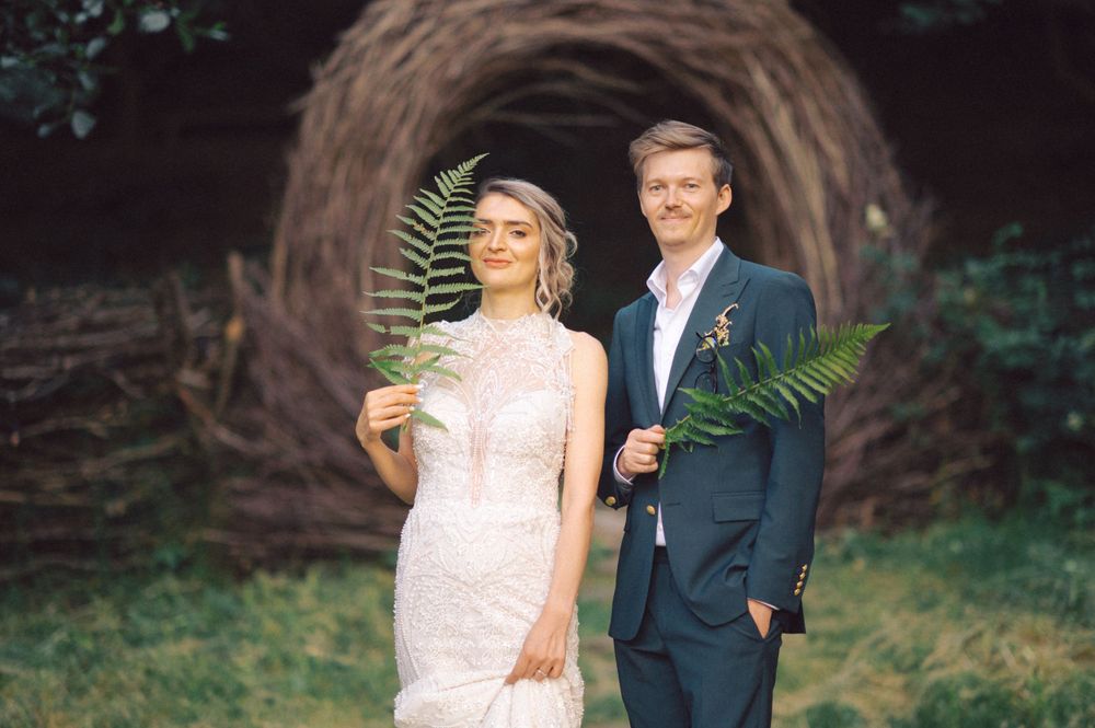 Wedding couple stands with fern leaves against a curved wooden art installation.
