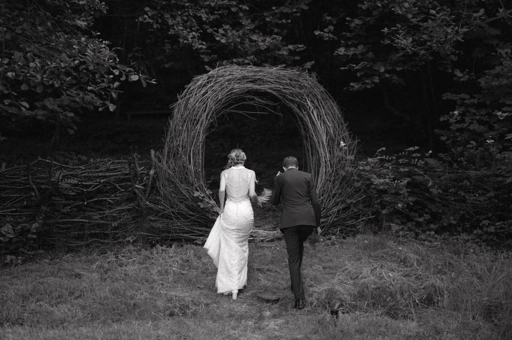 Black and white photo of couple walking towards large woven tunnel in garden.