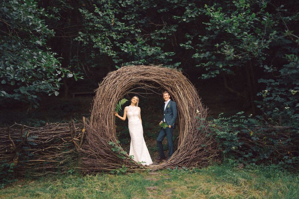 Wedding couple poses inside a large circular woven branch structure in a garden setting.