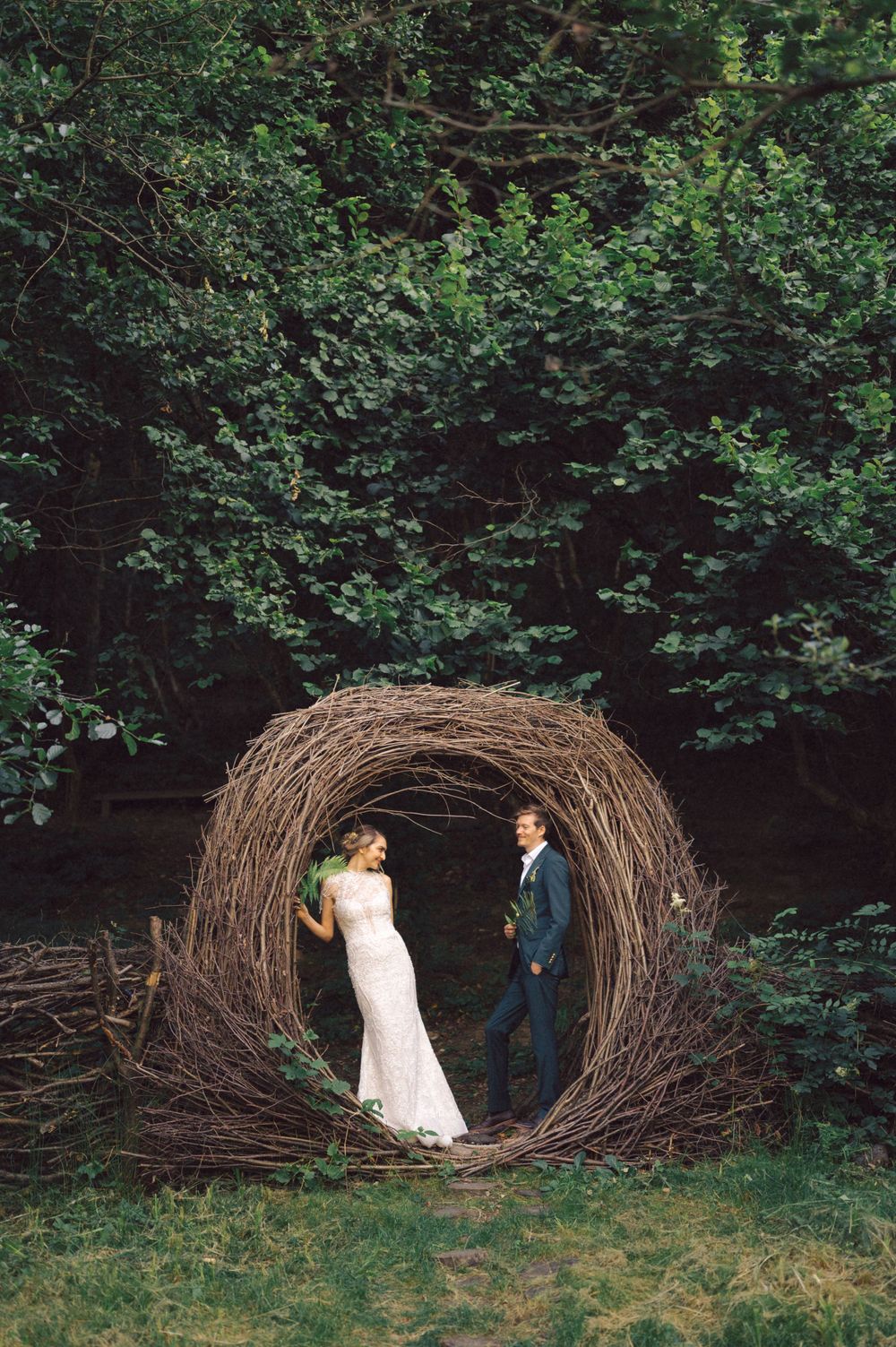 A couple standing inside a large circular nest-like structure made of branches in a woodland setting.