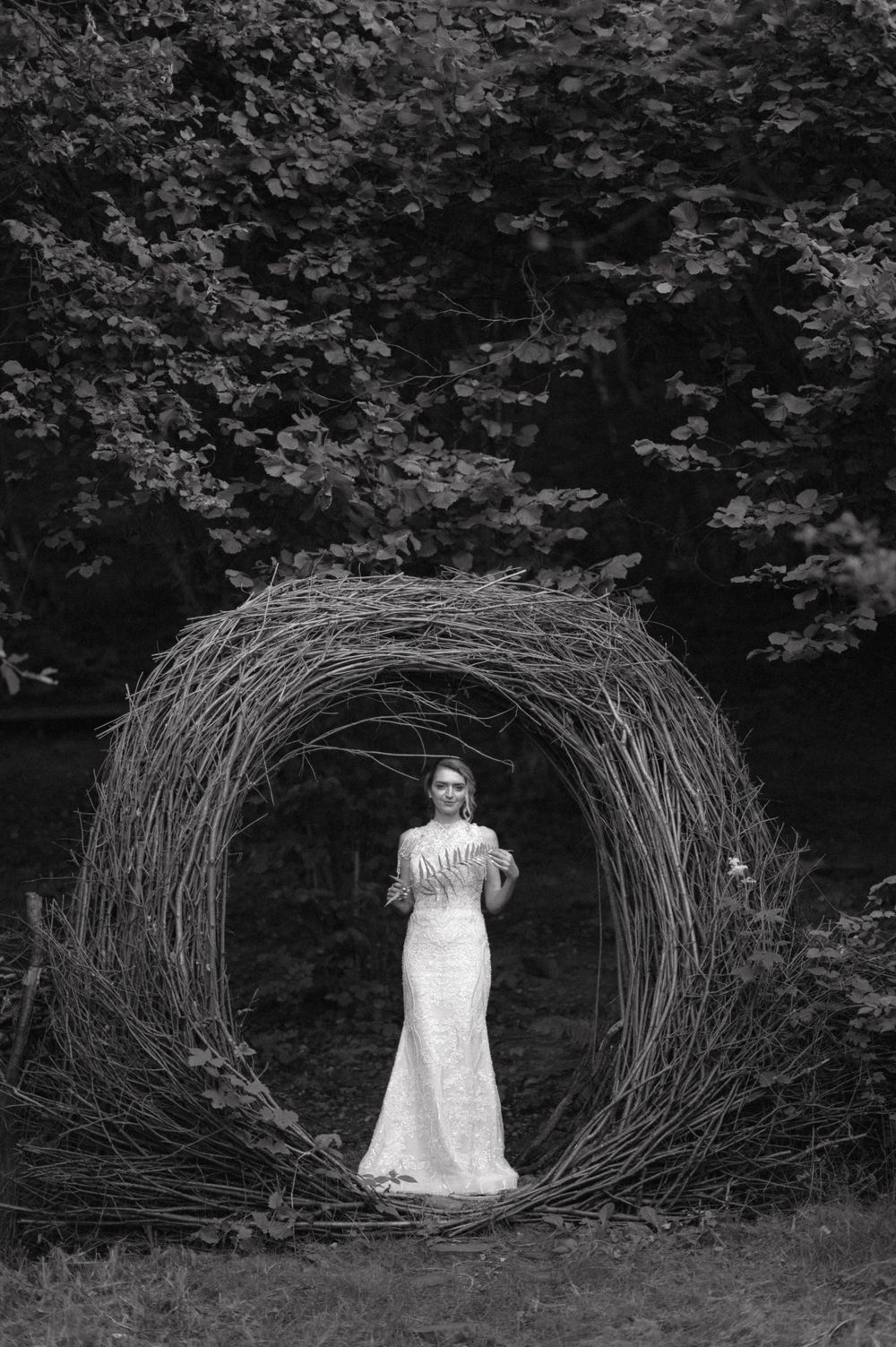 A figure in a white wedding gown poses in a circular woven branch archway against dark foliage.