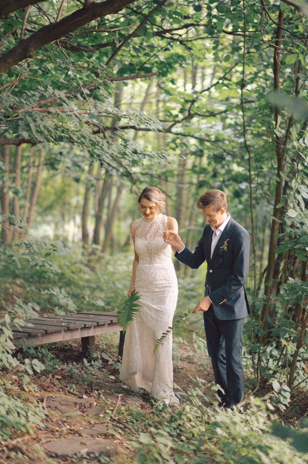 A couple walking together on a wooded path, with the person in white holding up their dress.