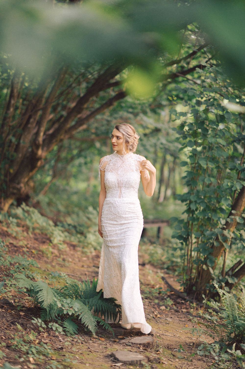 A figure in an elegant white lace dress poses on a woodland trail surrounded by greenery.