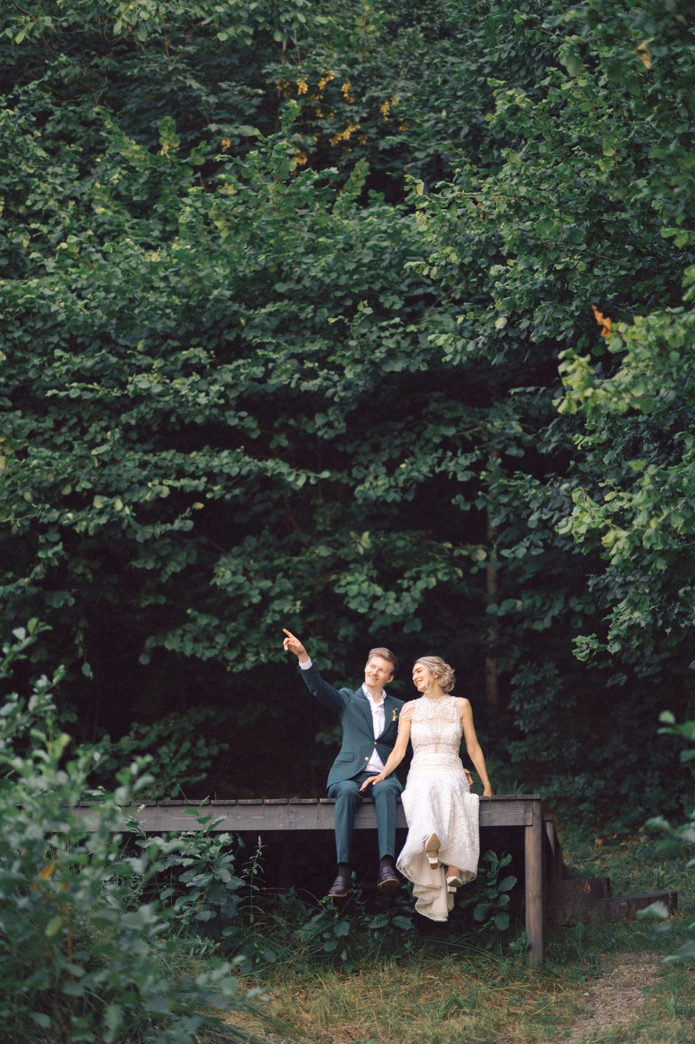 A couple sits intimately on a wooden bench surrounded by lush greenery in a romantic outdoor setting.