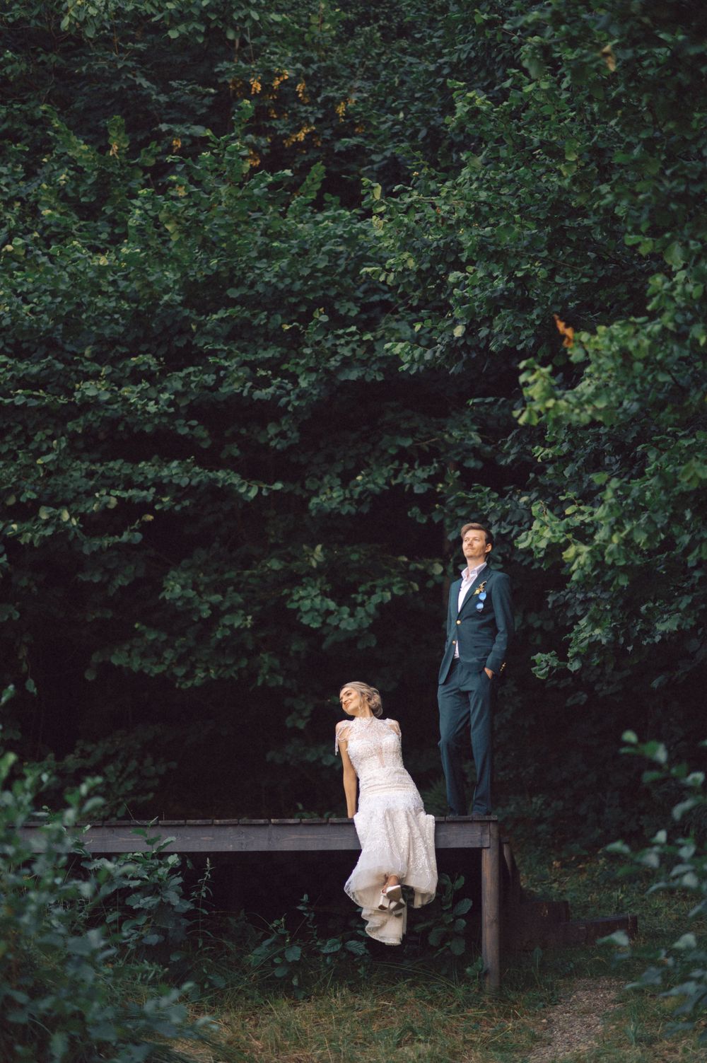 Two people pose playfully on a rustic wooden bench against a backdrop of dark forest foliage.