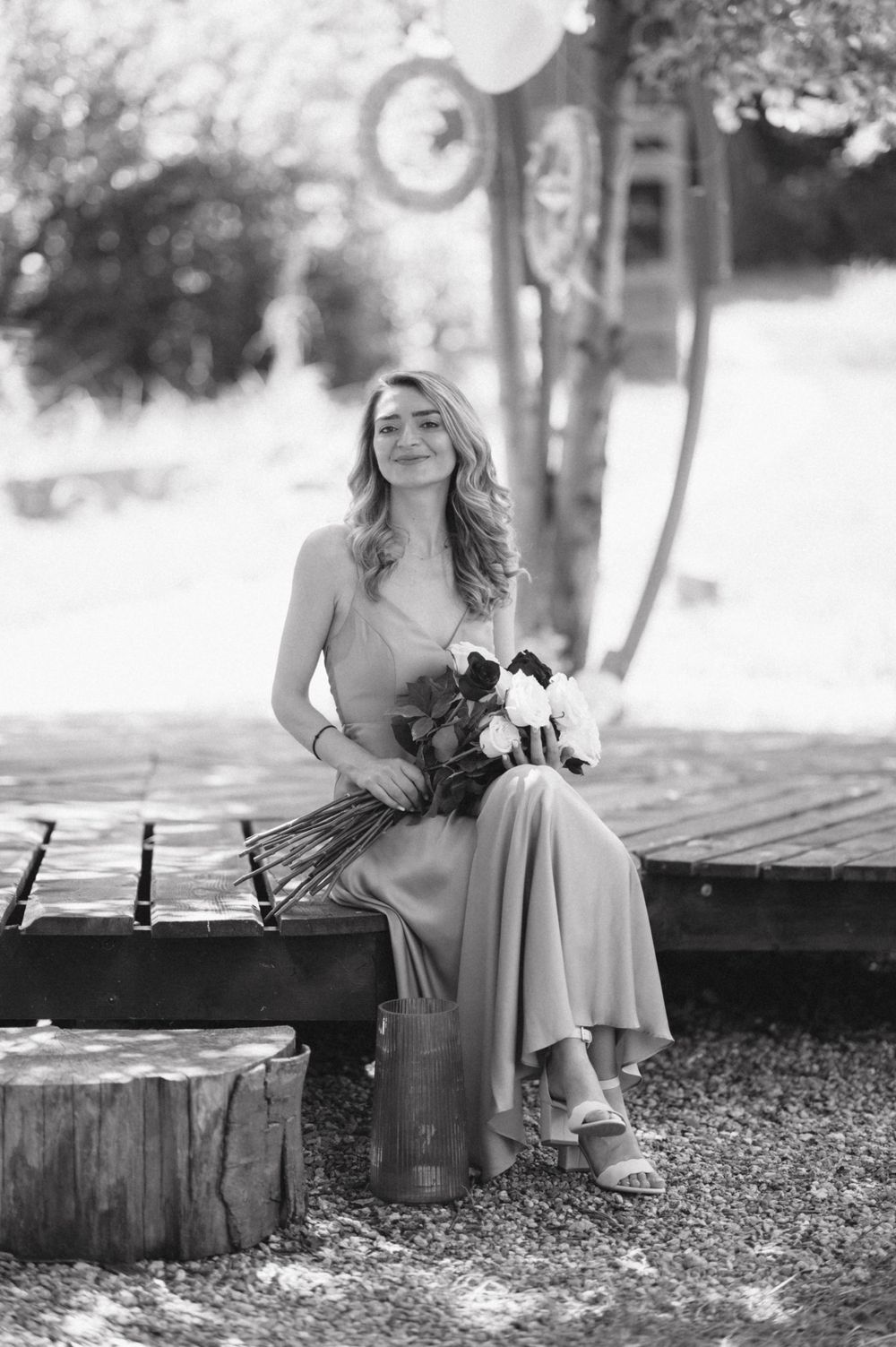 A woman in a flowing dress sits on a wooden platform holding a bouquet in black and white photography.