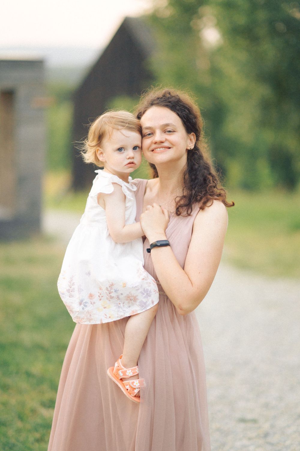 A woman in a pink dress holds a small child in a white floral dress outdoors.