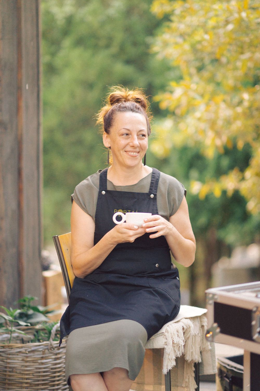 Person in overalls sits on rustic wooden bench holding a coffee cup outdoors.