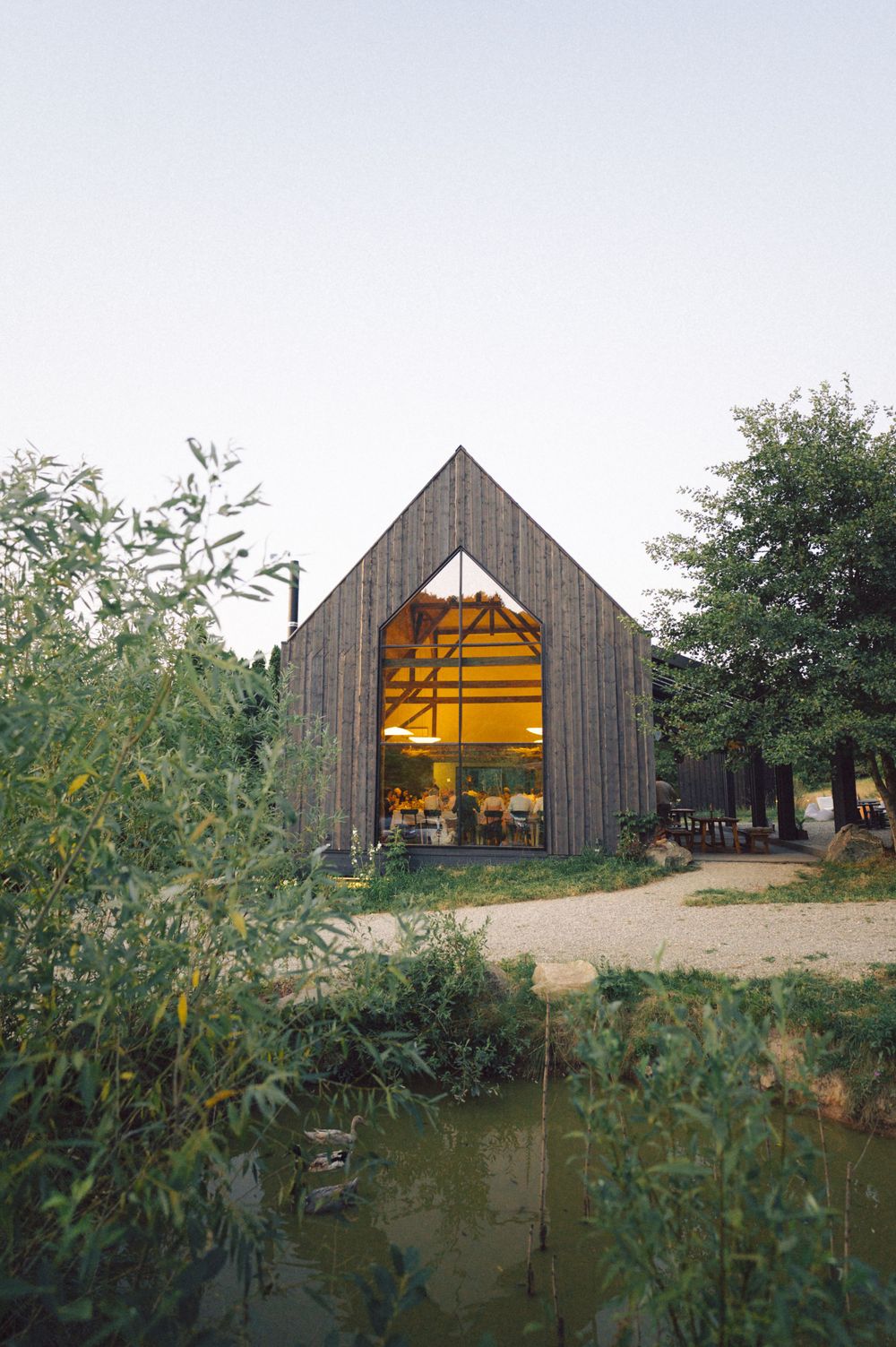 Illuminated wooden barn-style building with large glass doors reflecting warm light near a pond at dusk.