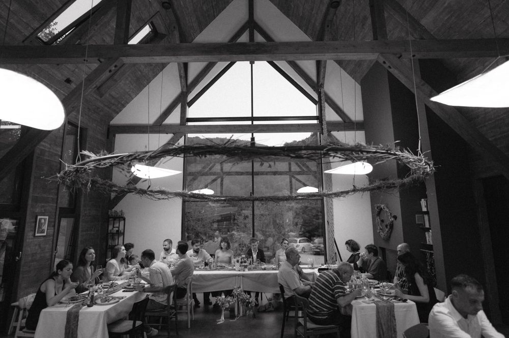 Black and white photo of a dining gathering inside a rustic barn with exposed beam ceiling.