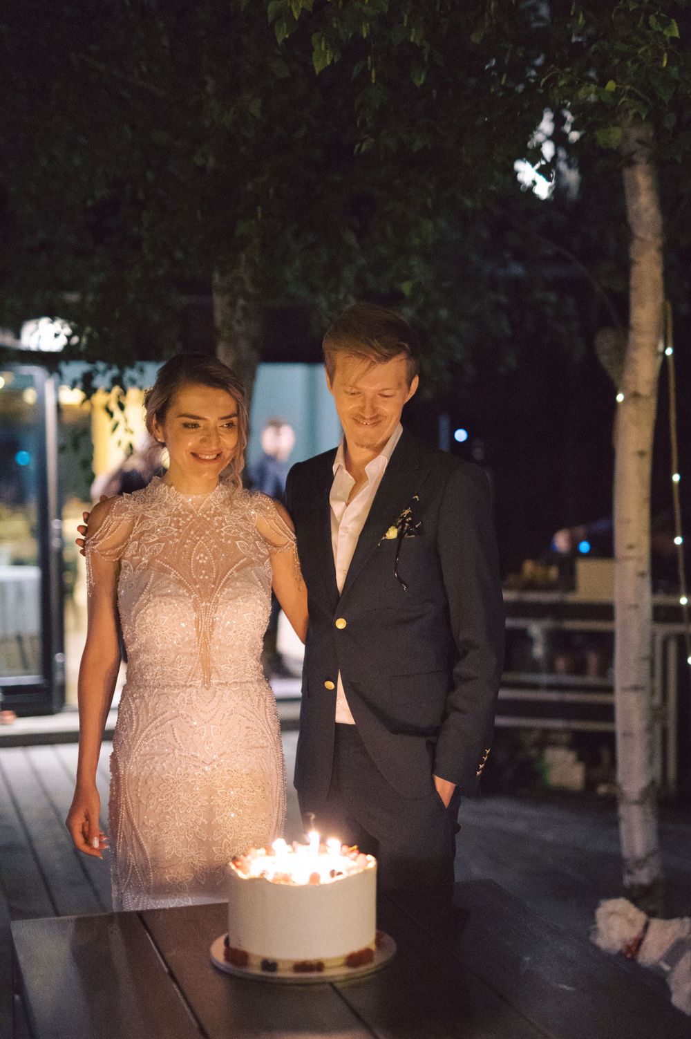 Evening celebration scene with a lit cake on a table at an outdoor venue.