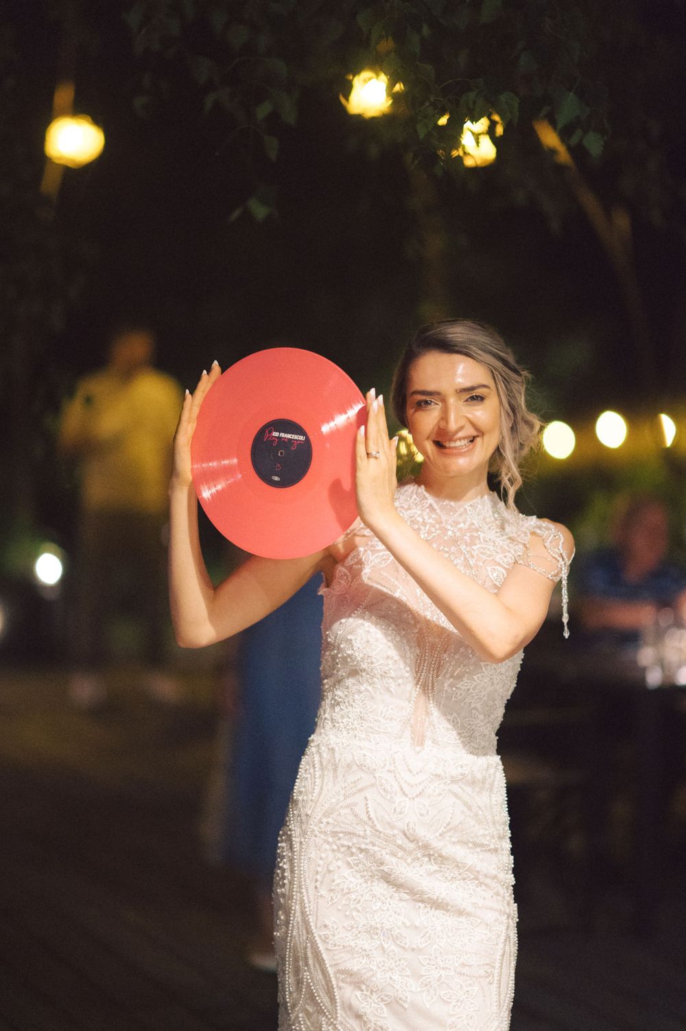 A bride in a beaded white dress holds up a red vinyl record at an evening reception.