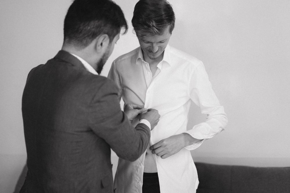 A candid black and white moment of two men helping with formal attire preparations.