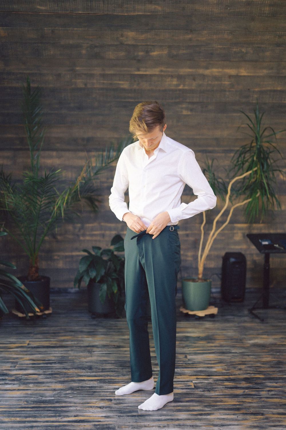 Person in white shirt and dark trousers adjusting clothing near potted plants.