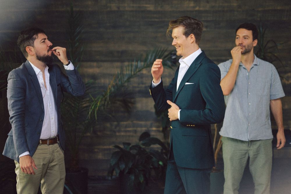 Three men in different attire share a candid moment against a rustic wooden wall backdrop.