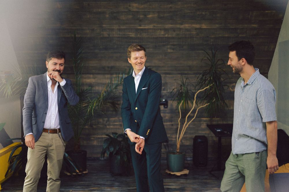 Casual group photo of three men laughing together in front of a wooden wall with plants.