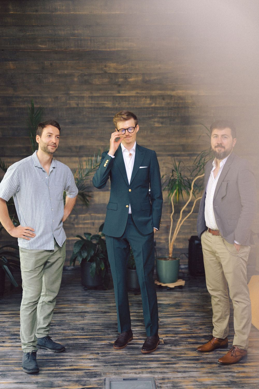 Three men in contrasting casual and formal wear pose against a wooden wall with potted plants.