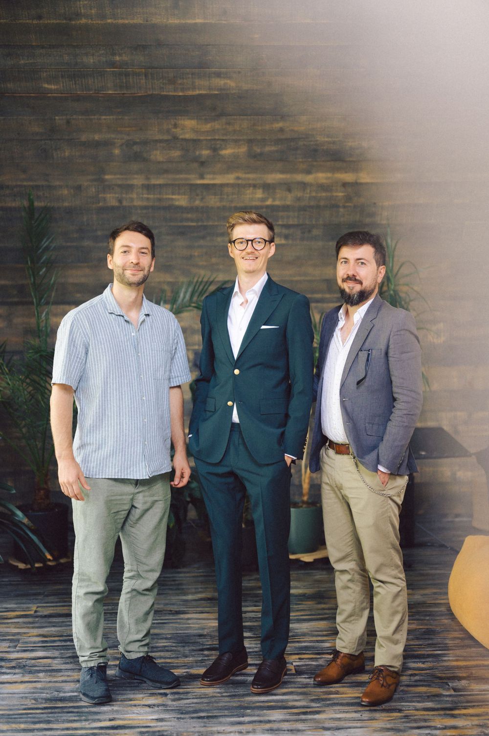 Group portrait of three men in varying styles of dress standing against rustic wood paneling.