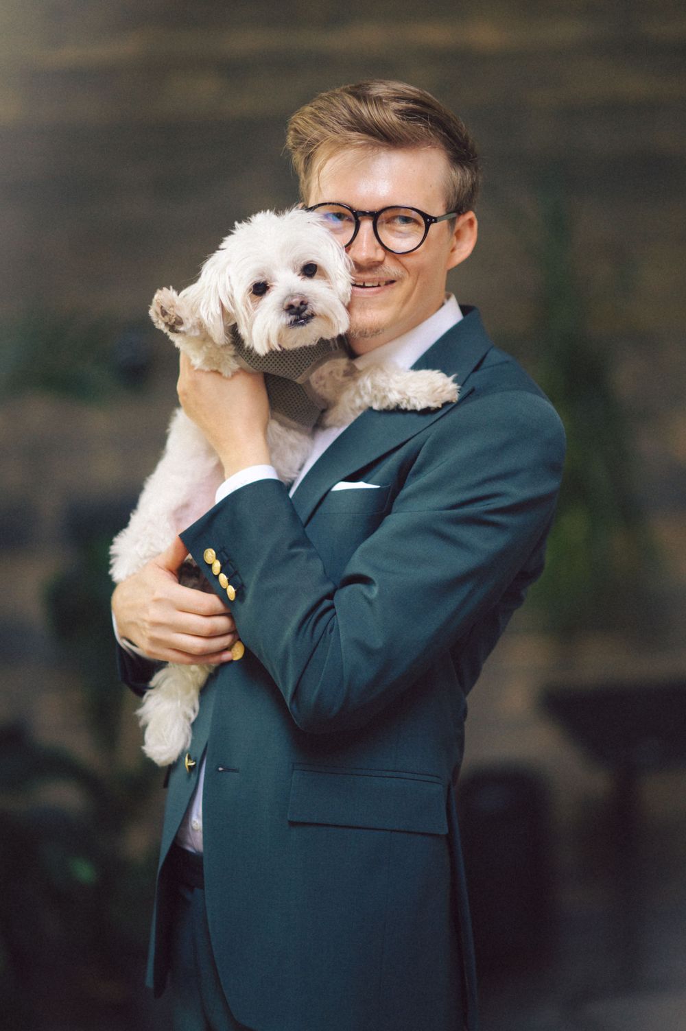 Person in teal suit jacket embraces small white dog against dark wooden backdrop.