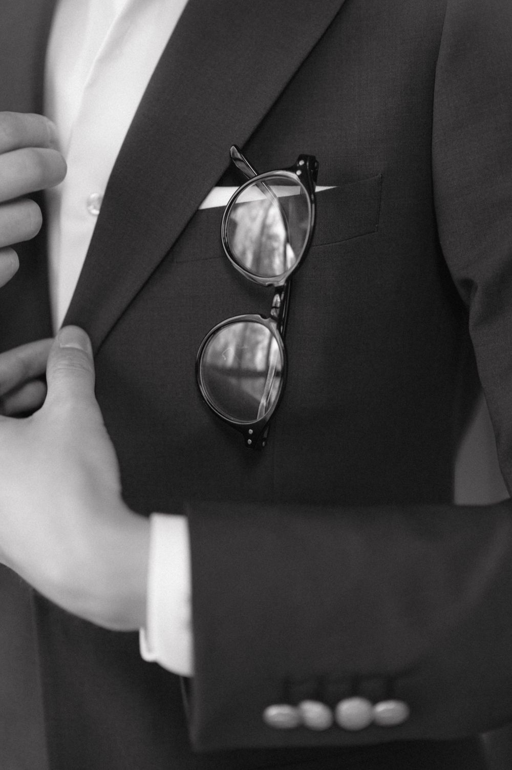 Black rimmed glasses rest elegantly on a dark suit jacket in a monochrome close-up shot.