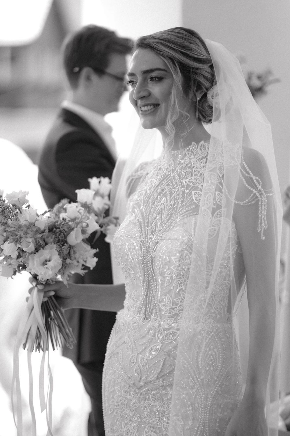 Black and white portrait of a bride in lace gown holding a floral bouquet.