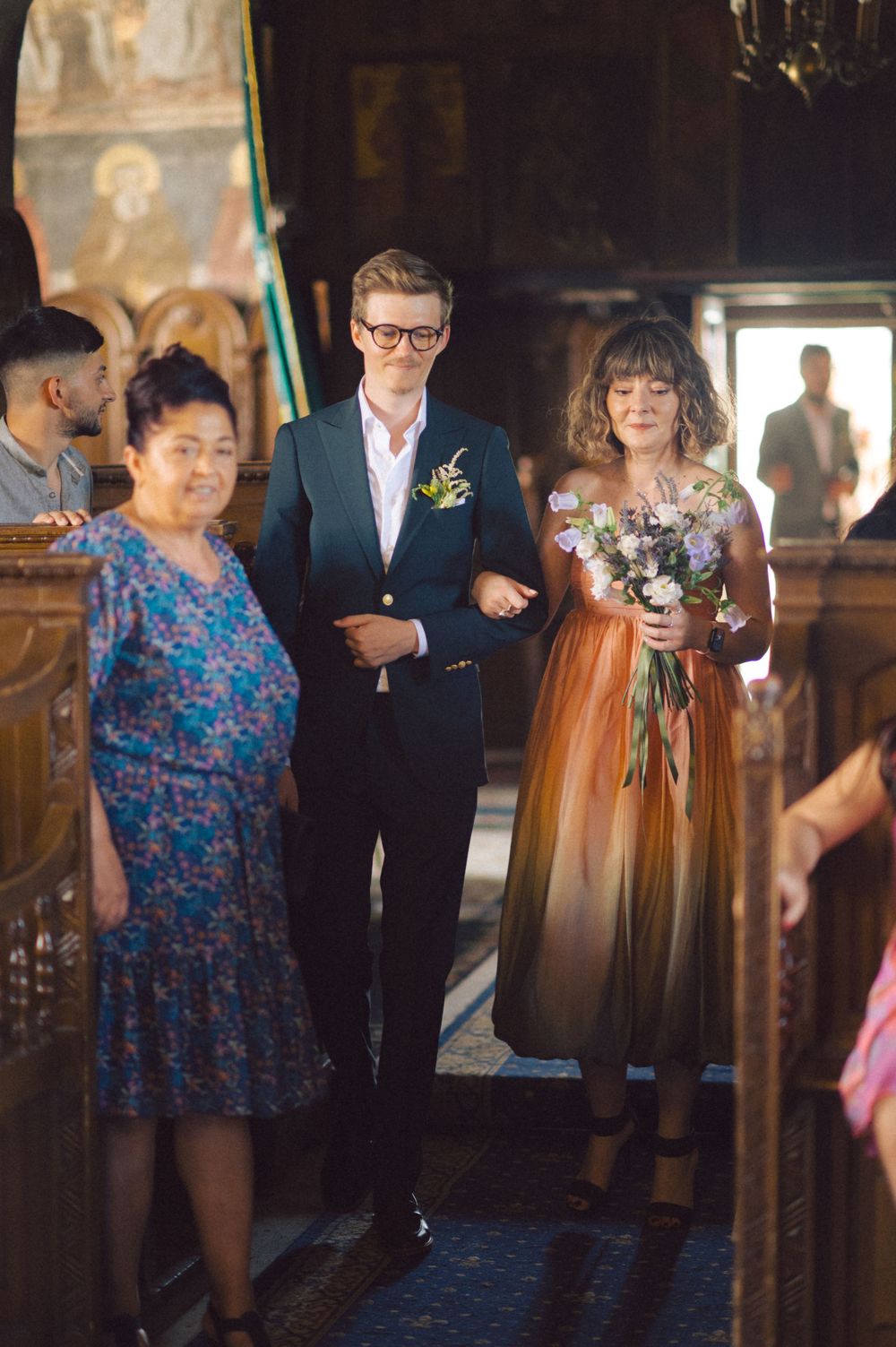 Wedding ceremony processional in a historic church interior with guests in colorful attire.