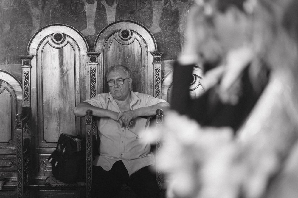A figure sits in an ornate wooden chair during a religious ceremony in black and white.