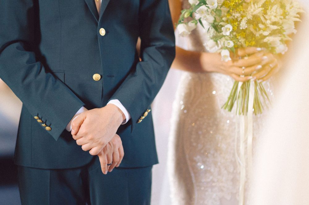 A formal navy suit and white wedding dress are shown in close-up during a wedding ceremony.