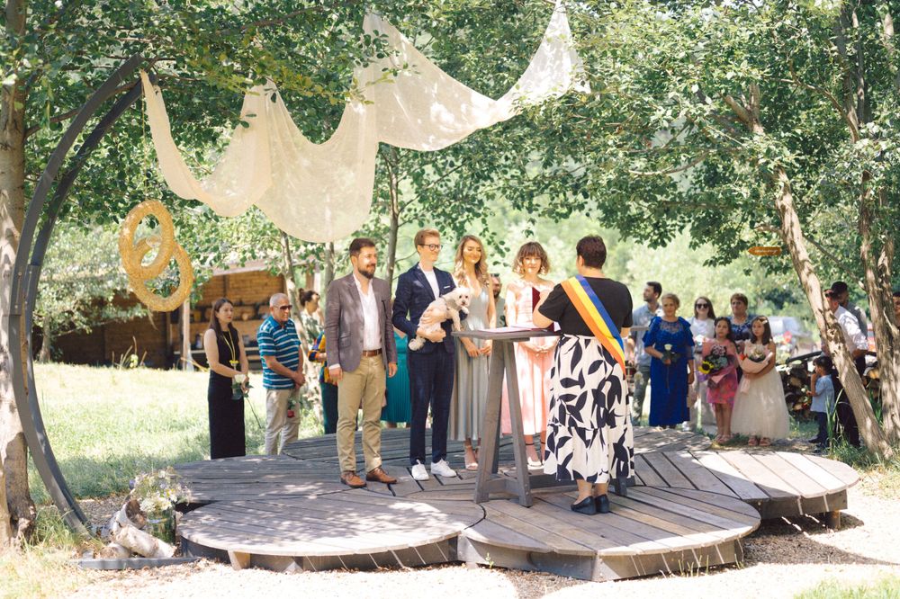 Wedding guests gather on a wooden platform under trees with flowing beige fabric decorations overhead for an outdoor ceremony.