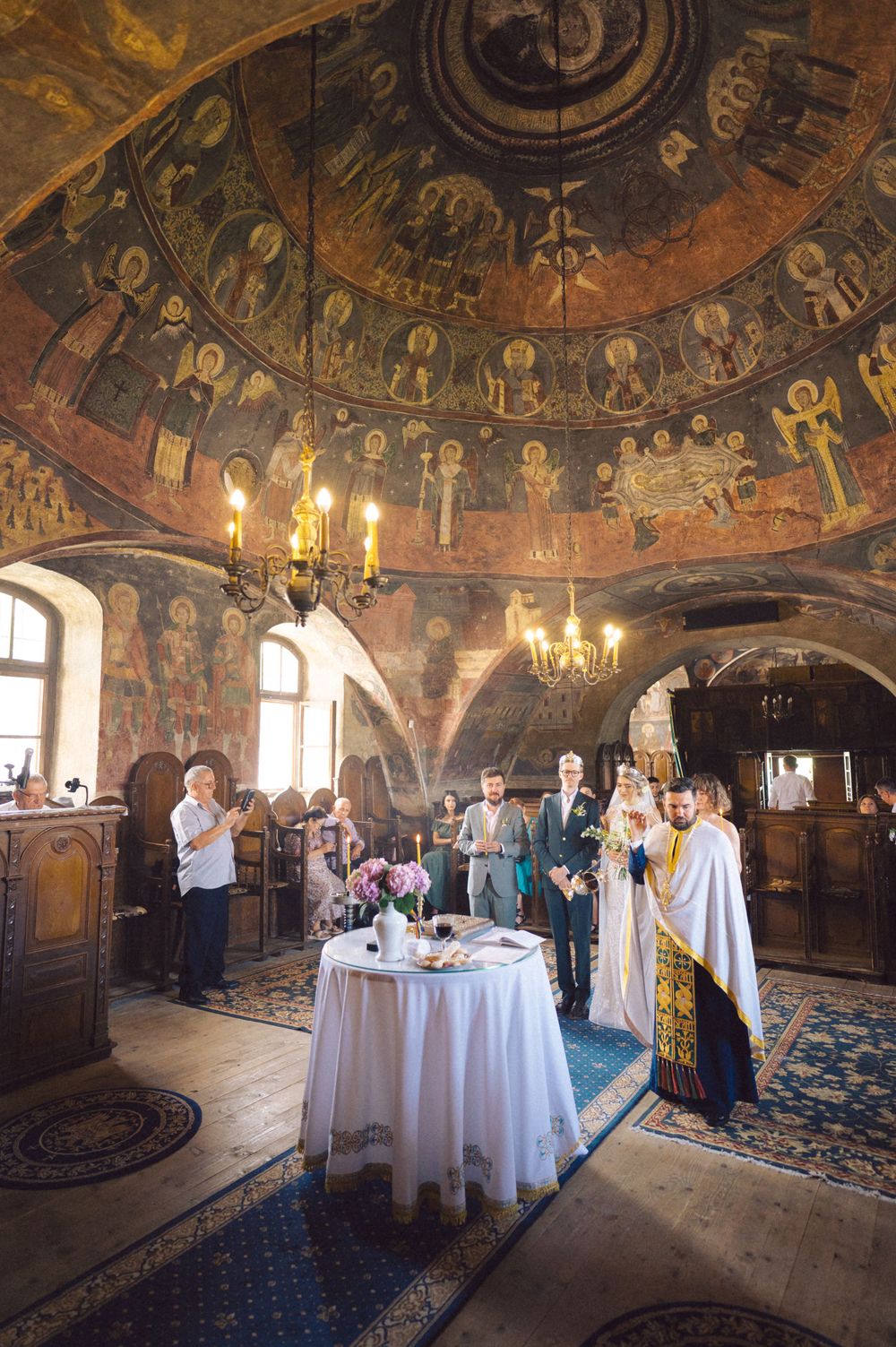An Orthodox church wedding ceremony beneath ornate ceiling frescoes and chandeliers.