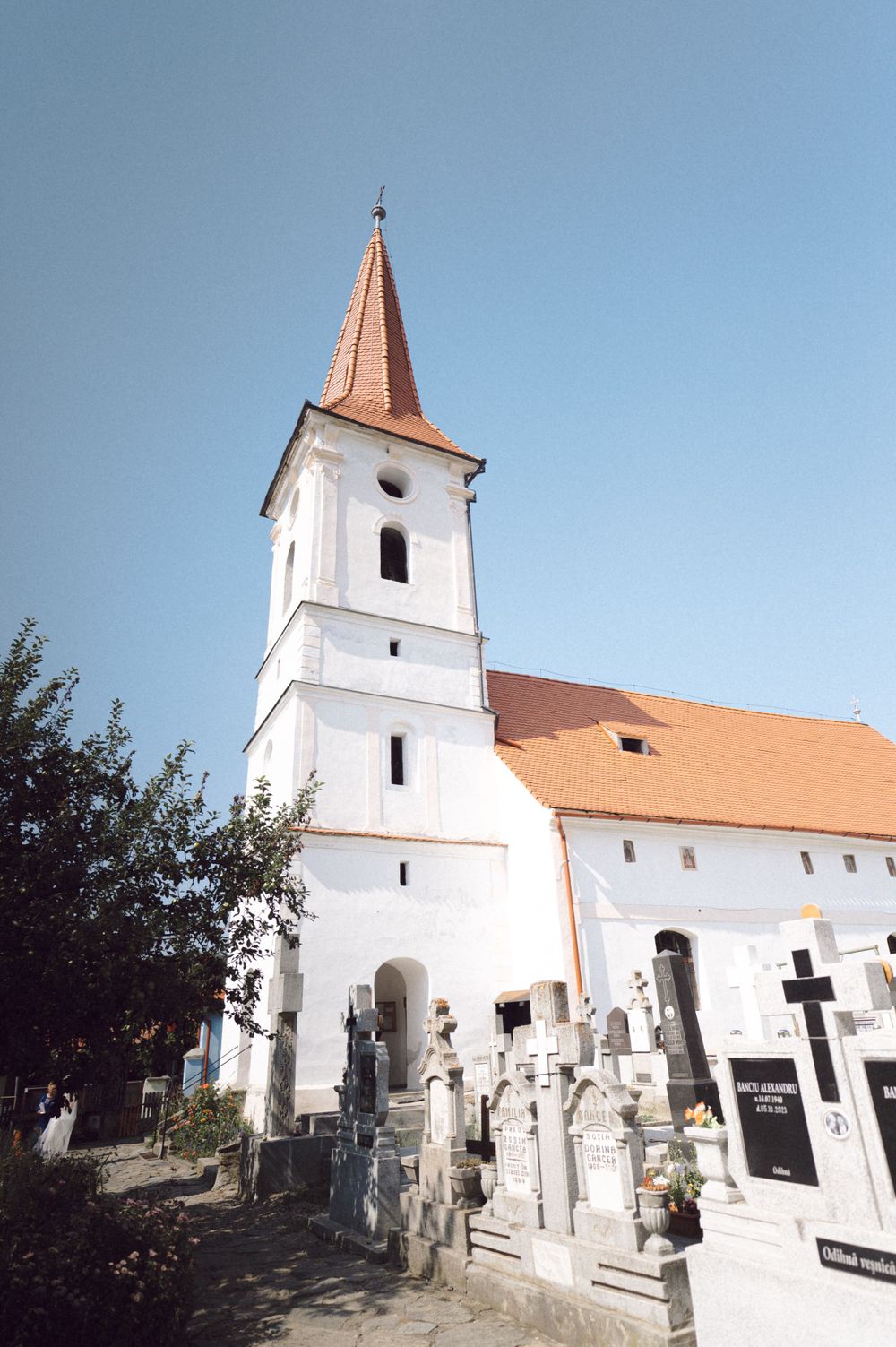 White church with tall spire and orange roof stands next to cemetery with stone monuments on a sunny day.