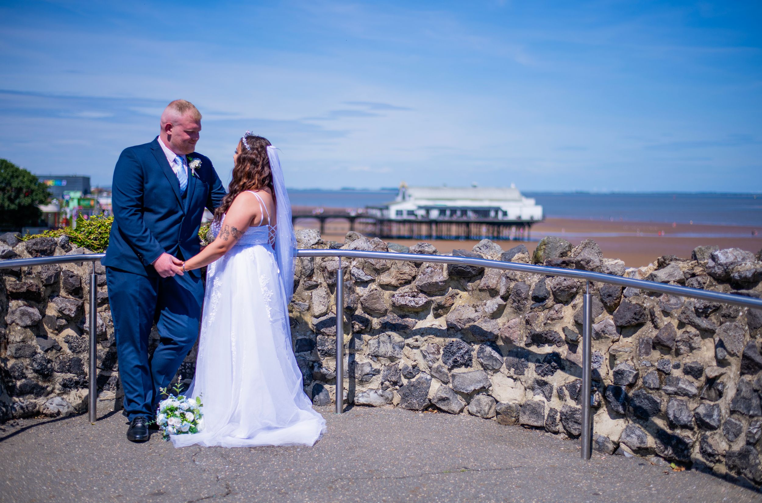 A couple poses by stone wall overlooking coastal view.