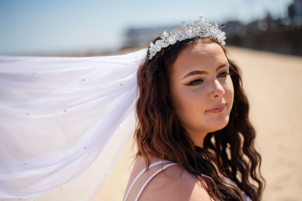 Flowing veil catches the breeze as sunlight illuminates a delicate crystal headpiece on a beach.