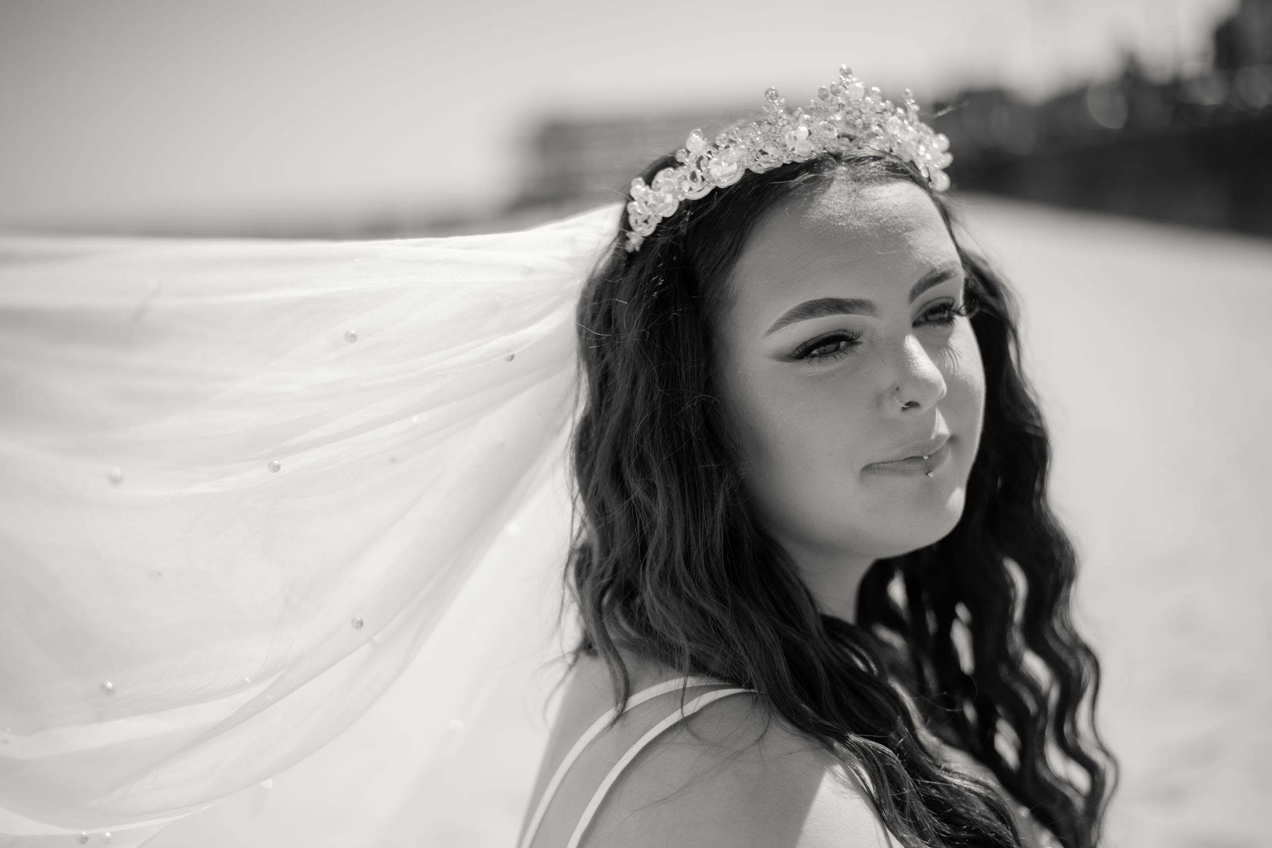 Black and white portrait showing a flowing veil and crystal headpiece against a coastal backdrop.