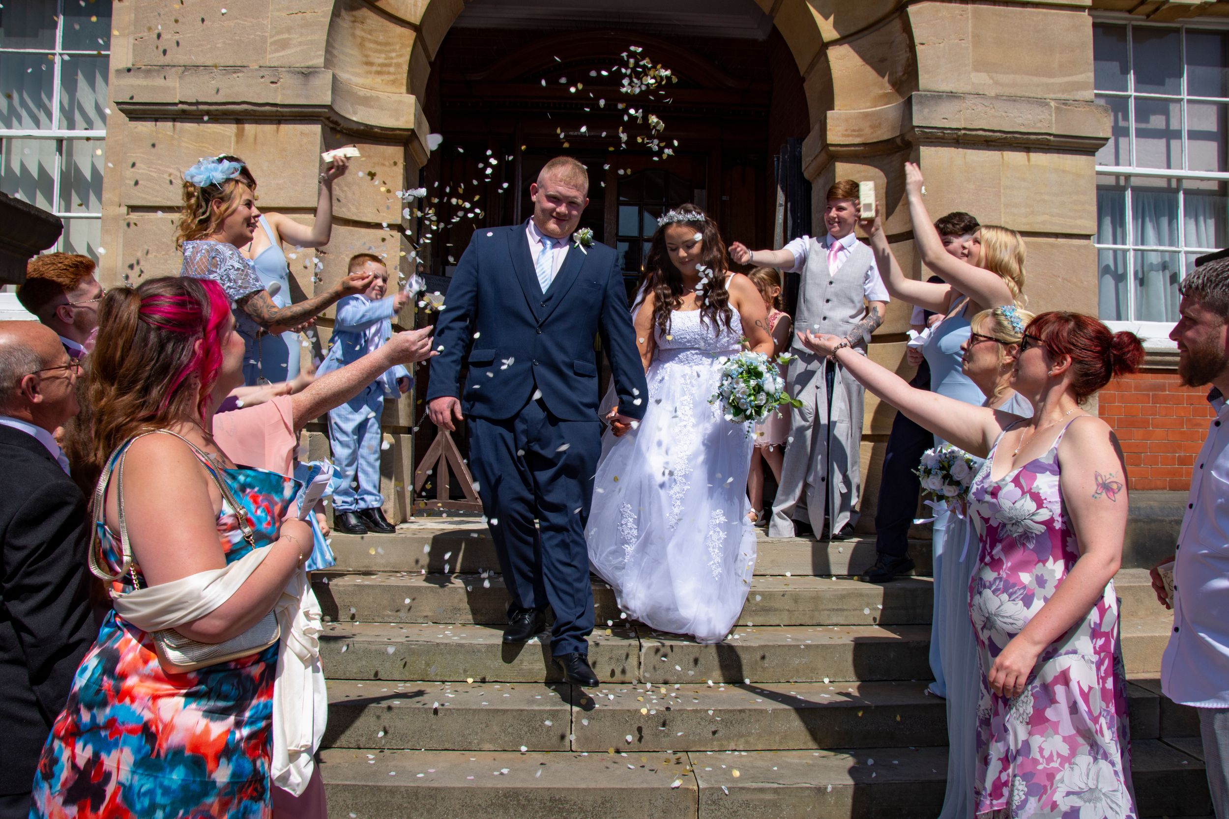 Wedding guests throw confetti as couple exits stone archway building.