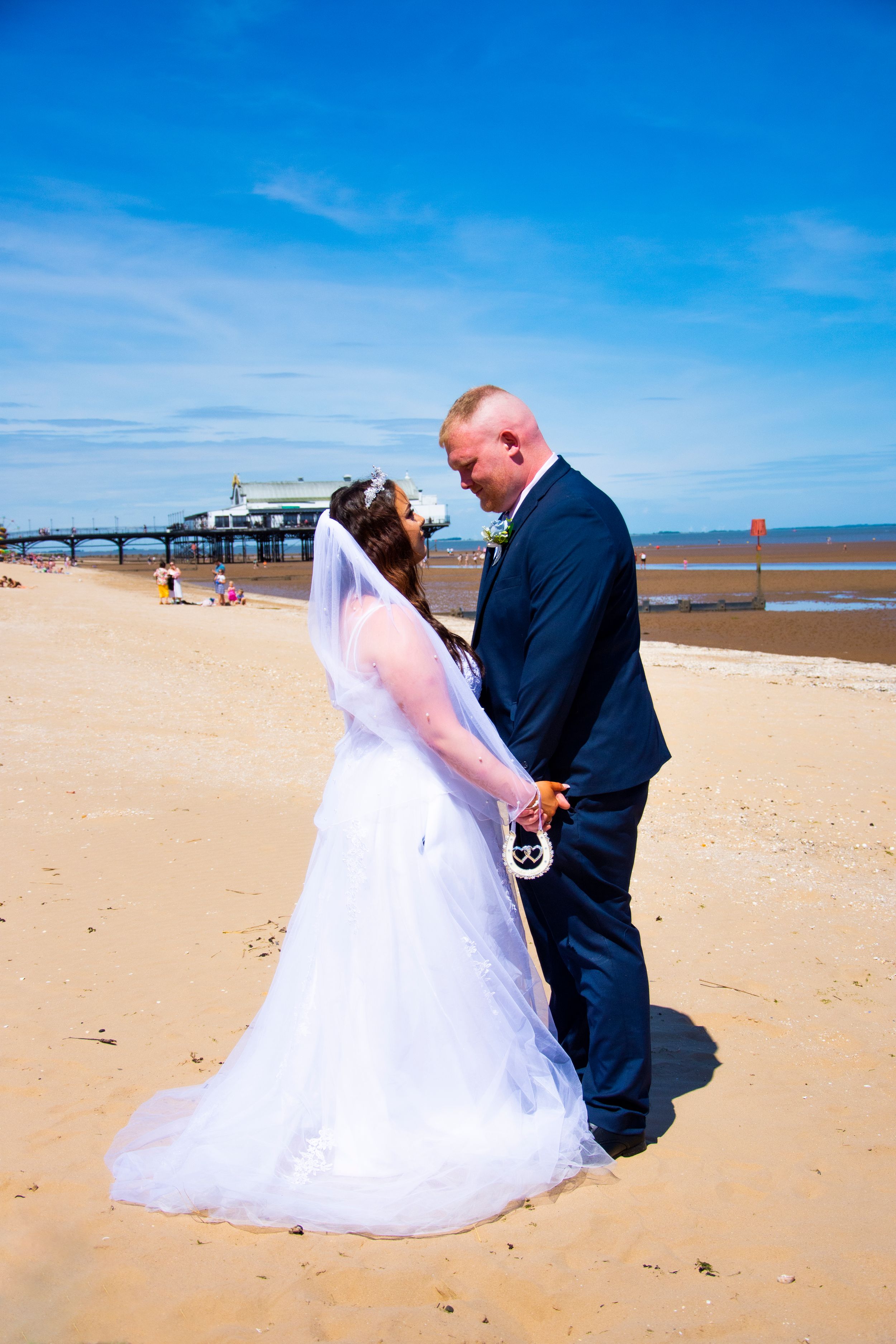 A couple in wedding attire standing on beach with pier in background.